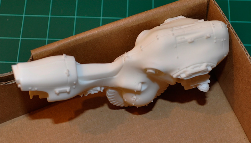 Unpainted white resin model parts with cylindrical and mechanical shapes resting inside a cardboard box, suggesting a work-in-progress modeling project.